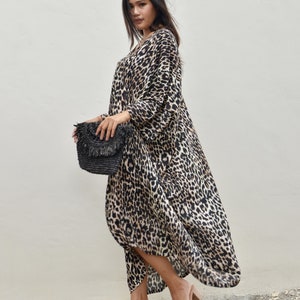 May include: A woman wearing a long, flowing leopard print dress with a black woven clutch bag and nude heeled sandals.