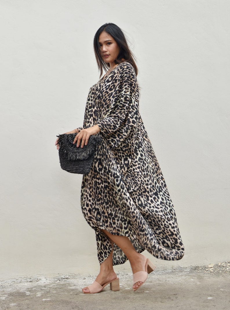 May include: A woman wearing a long, flowing leopard print dress with a black woven clutch bag and nude heeled sandals.