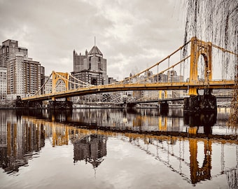 Könnte beinhalten: Eine gelbe Hängebrücke überspannt einen Fluss mit einer Skyline im Hintergrund. Die Brücke spiegelt sich im Wasser. Der Himmel ist bedeckt und grau.