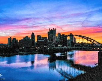 Könnte beinhalten: Ein Panoramablick auf eine Skyline bei Sonnenuntergang mit einer Brücke im Vordergrund. Der Himmel ist ein lebendiges Gemisch aus Rosa, Orange und Blau, wobei sich die Wolken im Wasser spiegeln. Die Stadtgebäude sind vor dem farbenfrohen Himmel silhouettiert.