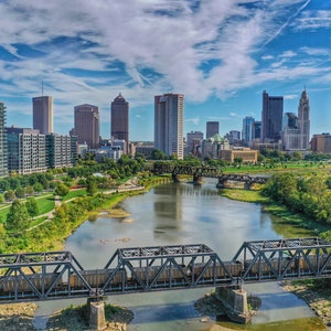 May include: An aerial view of a city skyline with a river and a bridge in the foreground. The city has many tall buildings, including skyscrapers. The river is wide and has a calm surface. The bridge is made of metal and has a dark gray color. The sky is blue and has some white clouds.