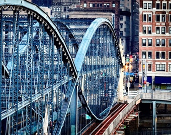 Könnte beinhalten: Eine Nahaufnahme einer Stahlbrücke mit zwei Bögen. Die Brücke ist hellblau gestrichen und hat ein komplexes Netzwerk aus Balken und Stützen. Die Brücke verläuft über eine Straße und im Hintergrund befinden sich Gebäude.
