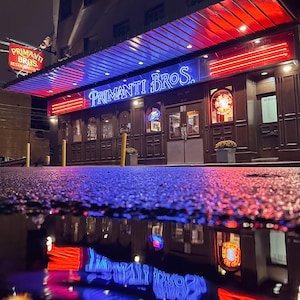 May include: A neon sign for Primanti Bros. Restaurant, with red and blue lights, is reflected in a puddle on the street.