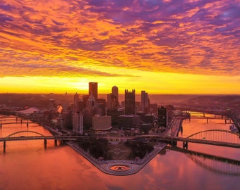 May include: An aerial view of a city skyline at sunrise with a vibrant orange and purple sky. The city is situated on a peninsula with two rivers flowing around it. Several bridges connect the city to the surrounding areas.