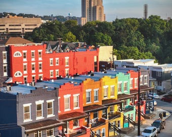 Peut inclure: Une vue en plongée d'une rangée de maisons de ville colorées dans des tons de bleu, rouge, orange, jaune et sarcelle, avec une haute tour gothique en pierre s'élevant en arrière-plan au-dessus d'une ligne d'arbres verts et d'une rue.