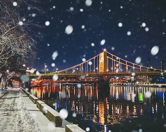 May include: A nighttime view of a bridge over a river with snow falling. The bridge is lit up with yellow lights and the river is reflecting the lights. There are trees on the left side of the image and a snowy path leading to the bridge.