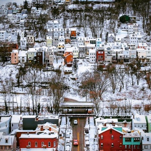 May include: A snowy winter scene with a street lined with houses and a bridge in the background. The houses are a mix of styles and colours, with some having snow-covered roofs. The street is empty, except for a few cars.