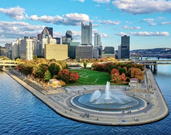 May include: An aerial view of a city skyline with a fountain in the foreground. The fountain is located on a small island in the middle of a river. The city skyline is in the background, with tall buildings and bridges.