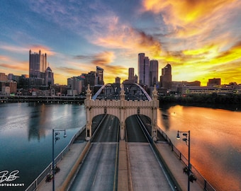 Könnte beinhalten: Eine Luftaufnahme einer Brücke über einen Fluss mit einer Skyline im Hintergrund. Die Brücke ist aus Beton und hat zwei Fahrspuren. Die Skyline der Stadt wird von der untergehenden Sonne beleuchtet, die einen goldenen Schein über die Szene wirft. Der Himmel ist eine Mischung aus Blau und Orange.