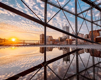 Könnte beinhalten: Eine Nahaufnahme einer Metallbrückenkonstruktion vor einem bewölkten Himmel und der untergehenden Sonne. Die Skyline der Stadt ist in der Ferne zu sehen, mit Reflexionen im Wasser darunter.