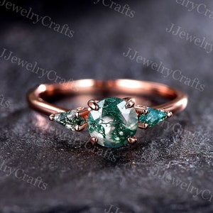 Op de afbeelding: Roze gouden ring met een groene mosagaat edelsteen en twee kleinere groene edelstenen. De ring ligt op een donkergrijze achtergrond.