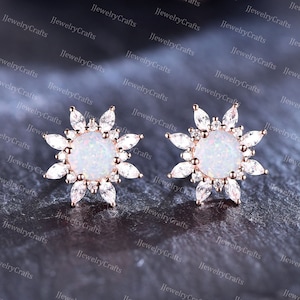May include: A pair of rose gold earrings with a white opal center stone and a halo of clear cubic zirconia stones. The earrings are shaped like a sunburst.
