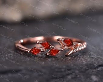 May include: A rose gold ring with a delicate branch design featuring red gemstones and small clear stones. The ring is set on a dark gray surface.