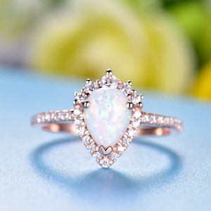 May include: Rose gold ring featuring a pear-shaped opal center stone, surrounded by a halo of small, clear gemstones. The band is also adorned with small gemstones. The ring is set against a light blue background.