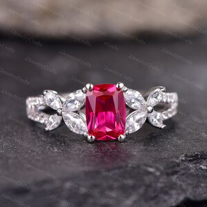 May include: A silver ring featuring a large, rectangular, vibrant red gemstone as the centerpiece. The ring is adorned with white, marquise-cut stones arranged in a floral pattern on either side of the main stone. The ring is set against a dark background.