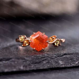 May include: A rose gold ring featuring a hexagonal orange gemstone as the centerpiece. The ring is adorned with leaf-like accents and smaller yellow-green gemstones on either side, creating a nature-inspired design. The ring is set on a dark, textured surface.