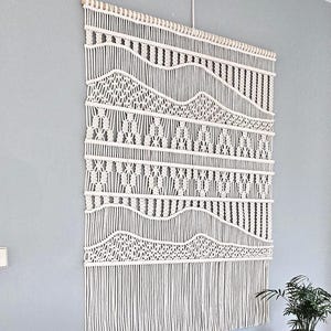 Op de afbeelding: Witte macramé wandhanger met een geometrisch patroon en franjes. De hanger is gemaakt van katoenen koord en is ongeveer 76 cm breed en 102 cm lang.