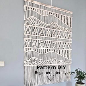 May include: A large off-white macramé wall hanging suspended from a wooden dowel, featuring intricate knotted geometric patterns and long fringe at the bottom. The text "Pattern DIY Beginners friendly" is printed in black with a heart icon.