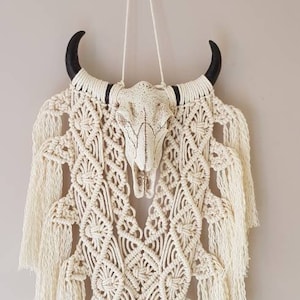 May include: A white macrame wall hanging with a white cow skull and black horns. The macrame is made with a knot pattern and has long fringe.