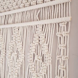 Puede incluir: Un gran tapiz de macram&eacute; de color blanco roto con un patr&oacute;n geom&eacute;trico. El tapiz est&aacute; hecho con cuerda de algod&oacute;n natural y tiene una varilla de madera en la parte superior. Una peque&ntilde;a etiqueta de madera con el texto "COEDANDMORE CREATIONS" est&aacute; unida al tapiz.