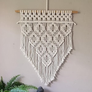 Op de afbeelding: Een witte macramé wandhanger met een geometrisch patroon, hangend aan een lichtbeige muur. Op de voorgrond staat een kleine groene plant en een grijs Boeddhabeeld.