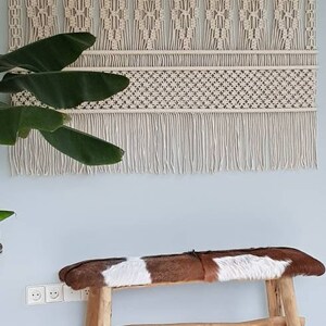 Puede incluir: Un tapiz de macram&eacute; blanco con un patr&oacute;n geom&eacute;trico y flecos. Un banco de madera con un asiento de piel de vaca marr&oacute;n y blanco est&aacute; en primer plano.
