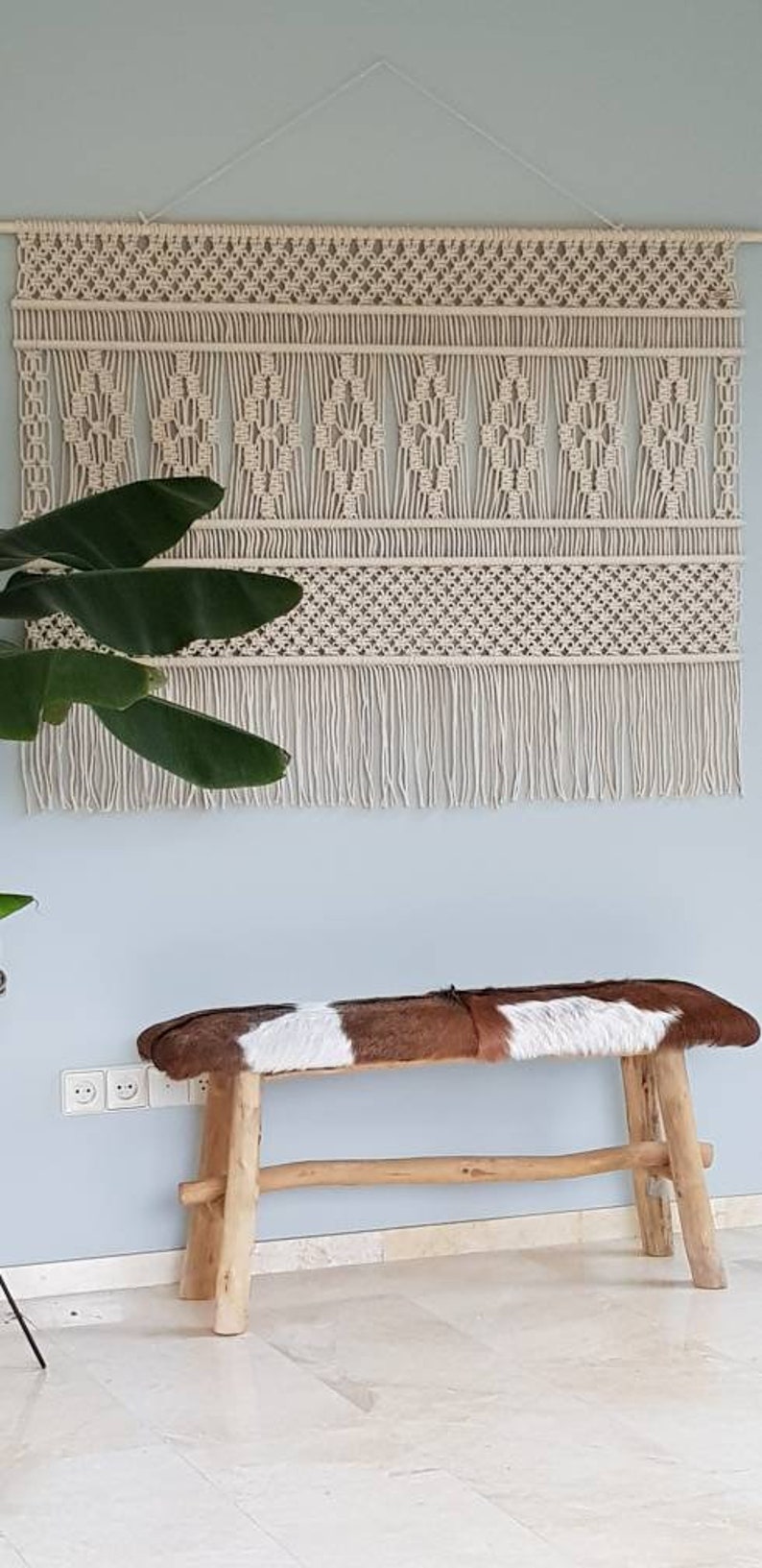 Puede incluir: Un tapiz de macram&eacute; blanco con un patr&oacute;n geom&eacute;trico y flecos. Un banco de madera con un asiento de piel de vaca marr&oacute;n y blanco est&aacute; en primer plano.