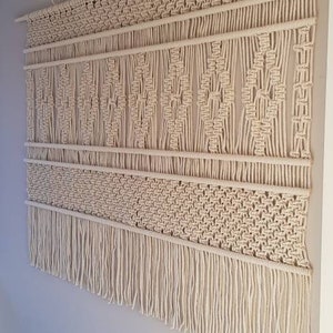 Puede incluir: Un gran tapiz de macram&eacute; de color blanco roto con un patr&oacute;n geom&eacute;trico y flecos. El tapiz est&aacute; hecho de cord&oacute;n de algod&oacute;n natural y est&aacute; colgado en una pared con una barra de madera.
