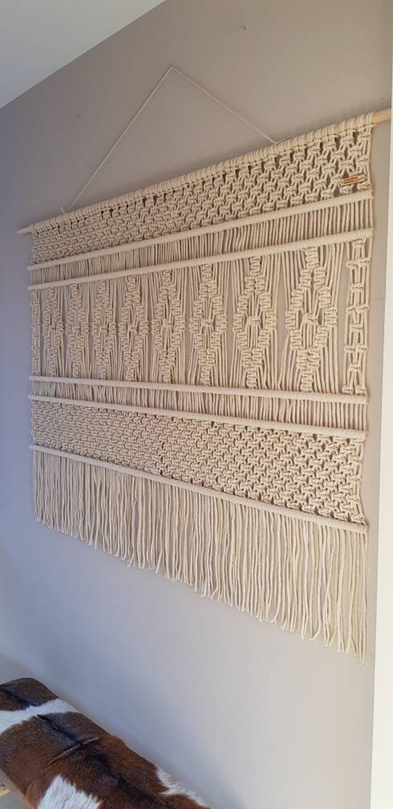 Puede incluir: Un gran tapiz de macram&eacute; de color blanco roto con un patr&oacute;n geom&eacute;trico y flecos. El tapiz est&aacute; hecho de cord&oacute;n de algod&oacute;n natural y est&aacute; colgado en una pared con una barra de madera.