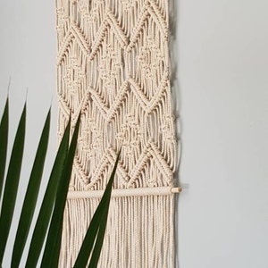 May include: A beige macrame wall hanging with a geometric diamond pattern and fringe. The hanging is made of natural cotton cord and has a wooden dowel at the top.