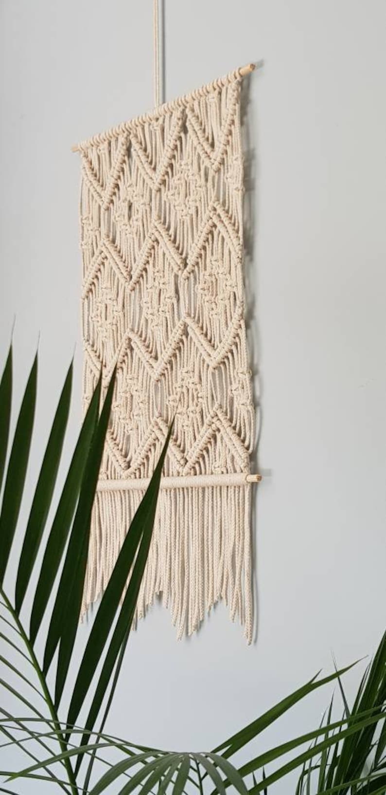 May include: A beige macrame wall hanging with a geometric diamond pattern and fringe. The hanging is made of natural cotton cord and has a wooden dowel at the top.