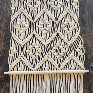 May include: A white macrame wall hanging with a geometric pattern and fringe. The hanging is made with natural cotton cord and is attached to a wooden dowel.
