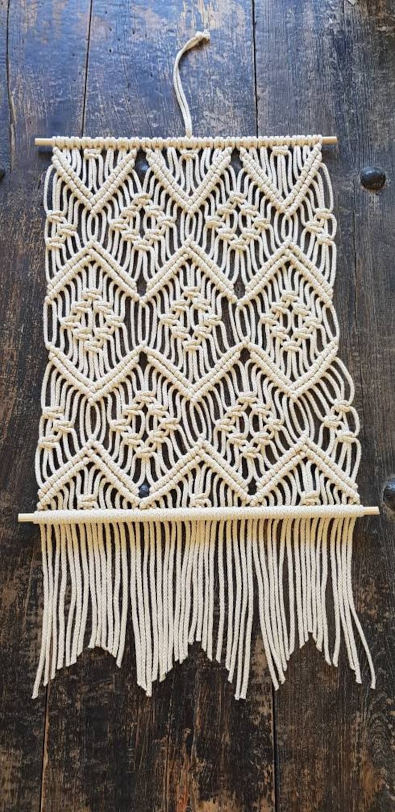 May include: A white macrame wall hanging with a geometric pattern and fringe. The hanging is made with natural cotton cord and is attached to a wooden dowel.