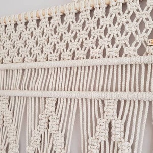Puede incluir: Un tapiz de macram&eacute; blanco con una barra de madera en la parte superior. El tapiz presenta un patr&oacute;n geom&eacute;trico con flecos en la parte inferior. El tapiz tiene una peque&ntilde;a etiqueta de madera con el texto "CREATIVECOTTAGECREATIVES" en ella.