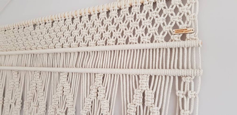 Puede incluir: Un tapiz de macram&eacute; blanco con una barra de madera en la parte superior. El tapiz presenta un patr&oacute;n geom&eacute;trico con flecos en la parte inferior. El tapiz tiene una peque&ntilde;a etiqueta de madera con el texto "CREATIVECOTTAGECREATIVES" en ella.