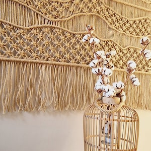 May include: A macrame wall hanging with a fringe border and a natural woven texture. A decorative bamboo cage lantern with a white cotton flower arrangement is in the foreground.