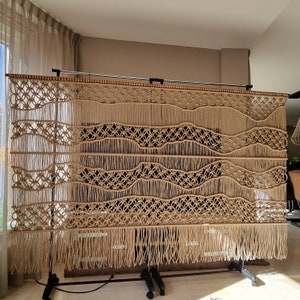 May include: A large macrame wall hanging with a woven pattern of waves and circles. The hanging is made of natural beige colored cord and has a fringe at the bottom.