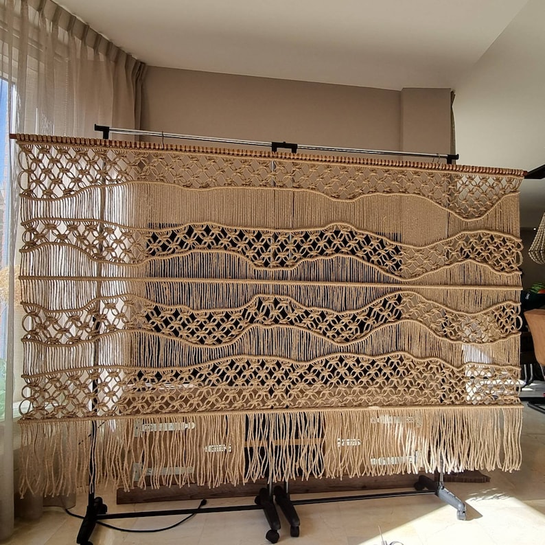 May include: A large macrame wall hanging with a woven pattern of waves and circles. The hanging is made of natural beige colored cord and has a fringe at the bottom.