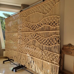 May include: A large macrame wall hanging with a wavy pattern and fringe. The hanging is made of natural beige colored cord and is mounted on a black metal stand.
