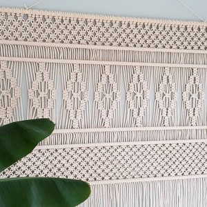 Puede incluir: Un gran tapiz de macram&eacute; de color blanco roto con un patr&oacute;n geom&eacute;trico. El tapiz est&aacute; hecho de cord&oacute;n de algod&oacute;n natural y tiene una franja en la parte inferior.