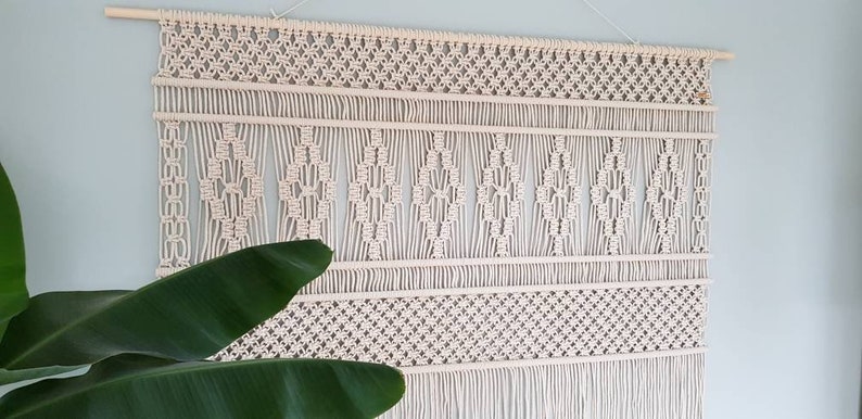 Puede incluir: Un gran tapiz de macram&eacute; de color blanco roto con un patr&oacute;n geom&eacute;trico. El tapiz est&aacute; hecho de cord&oacute;n de algod&oacute;n natural y tiene una franja en la parte inferior.