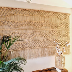 May include: A large macrame wall hanging with a woven pattern of circles and waves. The hanging has a fringe of tassels at the bottom and is made of natural beige colored rope.