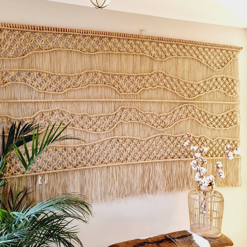 May include: A large macrame wall hanging with a woven pattern of circles and waves. The hanging has a fringe of tassels at the bottom and is made of natural beige colored rope.