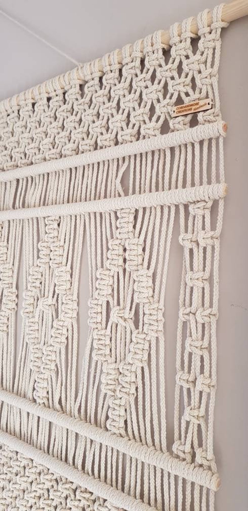 Puede incluir: Un gran tapiz de macram&eacute; de color blanco roto con un patr&oacute;n geom&eacute;trico. El tapiz est&aacute; hecho con cuerda de algod&oacute;n natural y tiene una varilla de madera en la parte superior. Una peque&ntilde;a etiqueta de madera con el texto "COEDANDMORE CREATIONS" est&aacute; unida al tapiz.