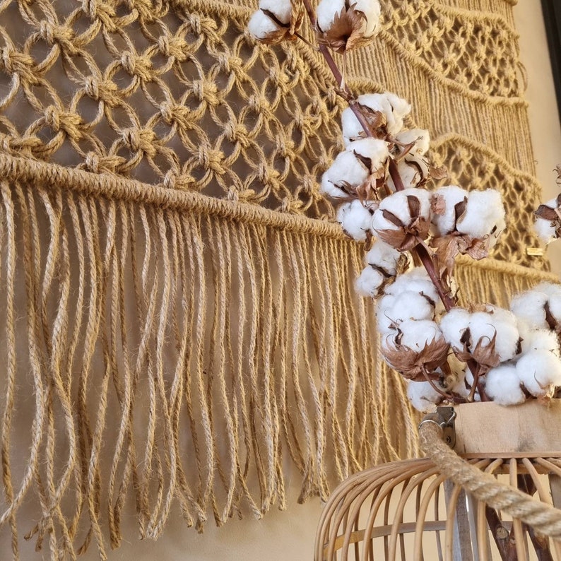 May include: A macrame wall hanging with a fringe border and a natural brown color. The wall hanging is decorated with a cluster of white cotton bolls.