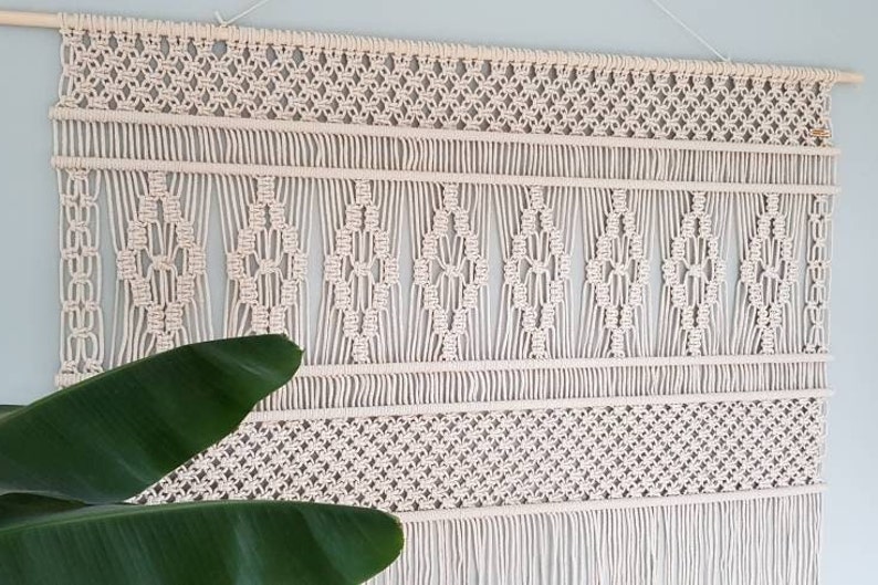 Puede incluir: Un gran tapiz de macram&eacute; de color blanco roto con un patr&oacute;n geom&eacute;trico. El tapiz est&aacute; hecho de cord&oacute;n de algod&oacute;n natural y tiene una franja en la parte inferior.