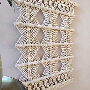 Peut inclure: Suspension murale en macramé crème avec un motif géométrique en losanges. La décoration murale est faite de corde de coton épaisse et tissée et est montée sur une cheville en bois naturel. Le design présente des nœuds complexes et un bord inférieur frangé.
