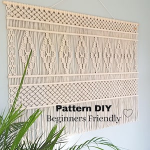 Op de afbeelding: Een grote, witte macramé wandhanger met een geometrisch patroon. De hanger is gemaakt van dik katoenen koord en heeft een franje aan de onderkant. De tekst "Pattern DIY Beginners Friendly" staat onder de hanger.