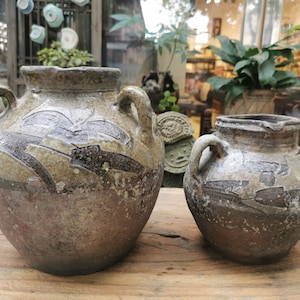 May include: Two brown ceramic jars with handles and a decorative design. The jars are sitting on a wooden surface.