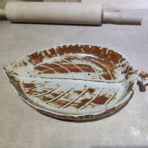 May include: A ceramic leaf-shaped serving dish with a brown glaze and a white background. The dish has a raised rim and a handle on one side.